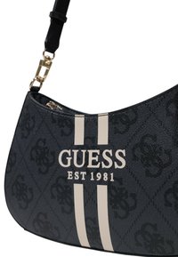 Sötétkék táska texturált külsővel, arany kiegészítőkkel, fehér csíkokkal és a "GUESS EST 1981" logóval hangsúlyosan megjelenítve.