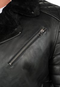 Zwarte leren jas met een schapenvachtkraag, een diagonale ritszak en een gewatteerde textuur op de schouders en bovenarmen.