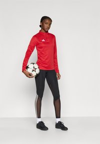 Κόκκινο αθλητικό μπλουζάκι μακρυμάνικο με ψηλό λαιμό και λευκό λογότυπο, σε συνδυασμό με μαύρο κολάν με διακοσμητικά από δίχτυ και λευκές ρίγες στο πλάι.