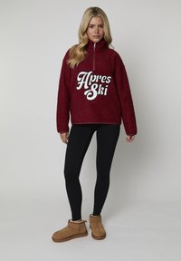 Burgundowy polarowy pulower z wysokim kołnierzem i zamkiem z przodu, z grafiką "Apres Ski" w kolorze białym. W połączeniu z czarnymi legginsami i beżowymi botkami.
