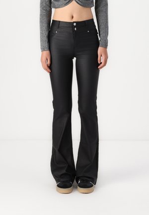 Kobieta w czarnych, rozkloszowanych spodniach z wysokim stanem, z przednimi kieszeniami i zapięciem na guziki, w zestawieniu z czarnymi trampkami z beżowymi podeszwami.