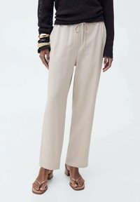 Beige, wijde broek gemaakt van zachte stof met een elastische tailleband en trekkoord. Gecombineerd met bruine sandalen en een zwarte gebreide top.