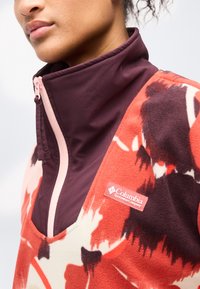 Κοντινό πλάνο ενός ατόμου που φοράει μπουφάν με φερμουάρ μπορντό και γιακά κάτω από ένα fleece της Columbia Sportswear με κόκκινες, μαύρες και λευκές σχέδια.