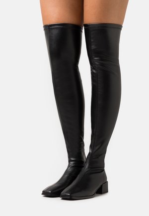 Bottes hautes noires en cuir slim, au-dessus du genou, avec des talons blocs bas, portées sur des jambes nues sur un fond blanc.
