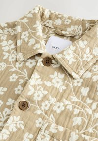 Cămașă bej cu un model floral, având o textură specială, cu flori albe, guler și două nasturi maro. Eticheta afișată spune "NEXT".