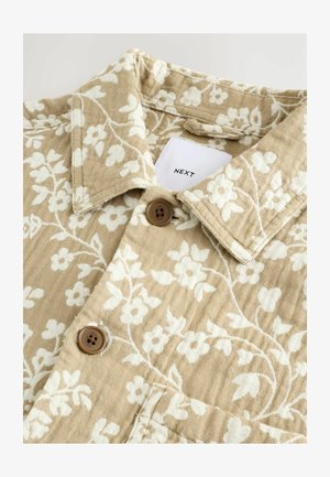 Beżowa koszula w kwiatowy wzór o teksturowanym wykończeniu, z białymi kwiatami, kołnierzykiem oraz dwoma brązowymi guzikami. Metka wyświetla napis "NEXT."