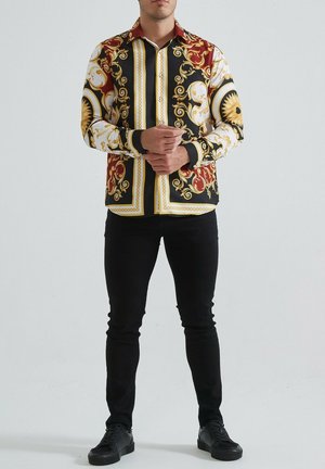 Chemise noire avec des motifs baroques dorés et rouges complexes. Manches longues, design à boutons, col contrasté, associée à un pantalon et des chaussures noirs.