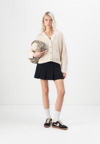 Beige gebreide cardigan met vier knopen, twee zakken, gecombineerd met een zwarte geplooide rok. Zwarte sneakers met witte accenten en grijze sokken.