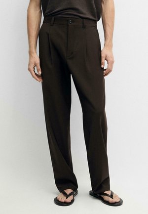 Mężczyzna ubrany w ciemnobrązowe plisowane spodnie i czarne klapki, stojący na jednolitym jasnym tle.