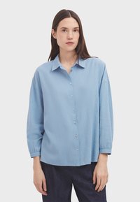 Camicia di cotone azzurro chiaro, con colletto, strisce verticali e maniche lunghe e ampie. Presenta piccoli bottoni e una vestibilità comoda.