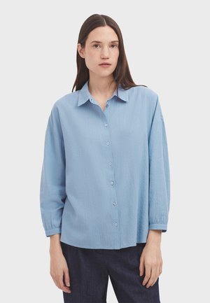Light blue button-up shirt with a collared neck, vertical stripes, and long, loose sleeves. Features small buttons and a relaxed fit.