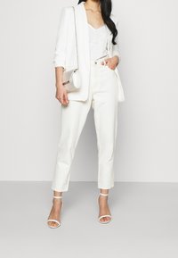 White blazer over a lace camisole, paired with cream-colored trousers. Accessorized with a white handbag and strappy white heels.