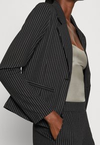 Czarny żakiet w paski o dopasowanym kroju, z białymi paskami, klapami i kieszenią z przodu, nałożony na jasnoszary top.