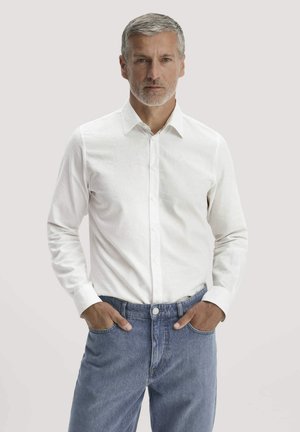 Mann mit grauen Haaren und Bart, der ein weißes Hemd mit Knöpfen und blaue Jeans trägt, steht mit den Händen in den Taschen vor einem einfachen Hintergrund.