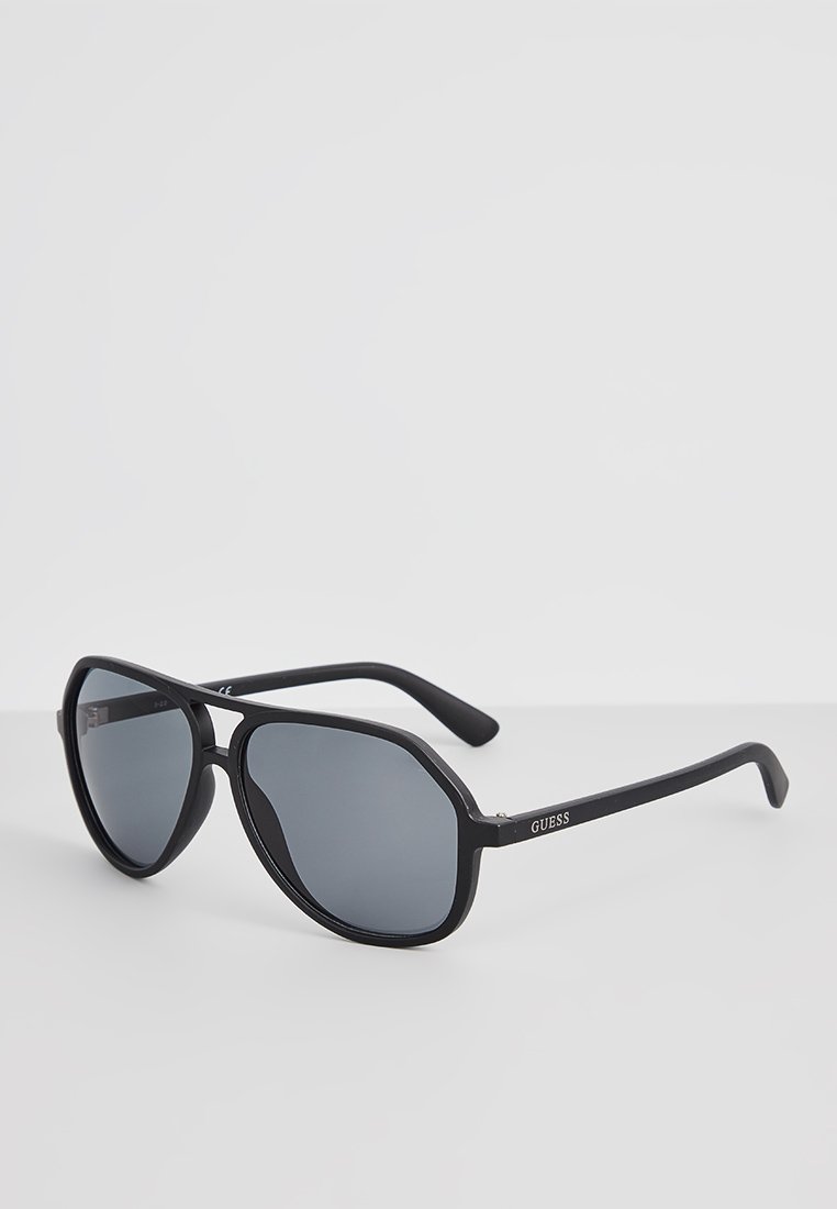 Occhiali da sole neri con lenti grigie, caratterizzati da un design arrotondato e una montatura spessa di colore nero opaco. Logo del brand visibile sulla stecca.
