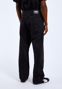 Black denim pants with a relaxed fit, featuring a logo patch on the back waistband, two rear pockets, and a smooth texture.