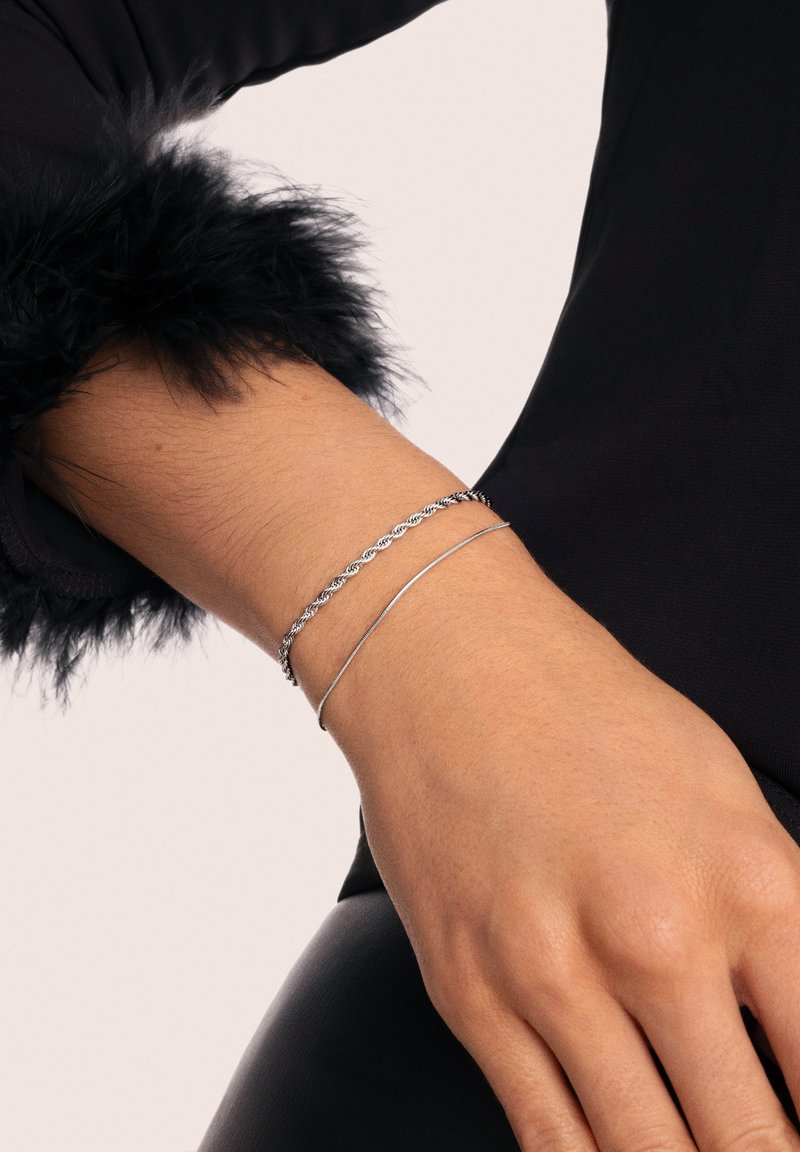 Brazaletes de plata llevados en una muñeca: uno retorcido, el otro liso y cilíndrico. La muñeca está parcialmente cubierta con piel sintética negra.