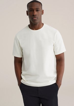 Wit T-shirt met korte mouwen, gestructureerde stof met een geribbelde afwerking, ronde hals, gecombineerd met donkere broek, handen in de zakken. Neutrale achtergrond.