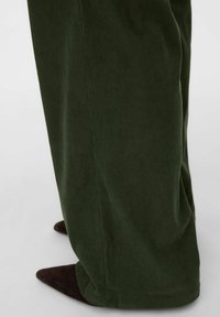 Szerokie, ciemnozielone spodnie sztruksowe o miękkiej fakturze, z pionowymi prążkami. U dołu częściowo widoczne brązowe buty w szpic.