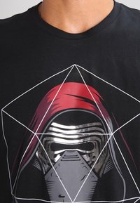 Černé tričko s designem kovové masky a červené kapuce překryté bílými geometrickými liniemi ve tvaru kostek.