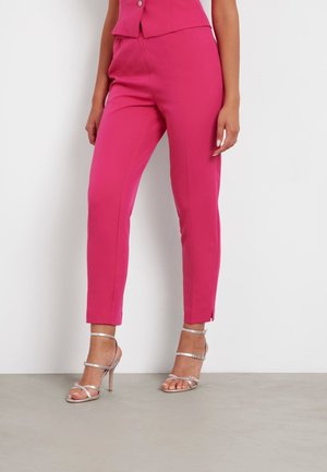 Pantalons ajustés roses avec une coupe fuselée, présentant des fentes sur le côté au niveau des chevilles, associés à des escarpins argentés à lanières sur un fond neutre.