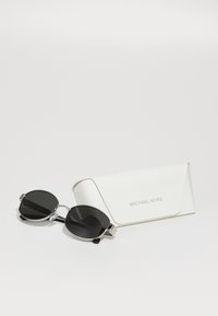 Ochelari de soare ovali negri cu accente argintii, însoțiți de un toc alb pe care este gravat numele brandului "MICHAEL KORS" în gri.