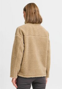 Veste en polaire beige avec un col haut, un tissu texturé et des épaules tombantes. Dotée d'un bas ourlé et de détails de couture simples.