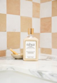 Flacon de revitalisant Rahua Classic avec bouchon doré sur un plan de travail en marbre contre un mur carrelé beige et blanc avec un petit bol et un bâton en bois.