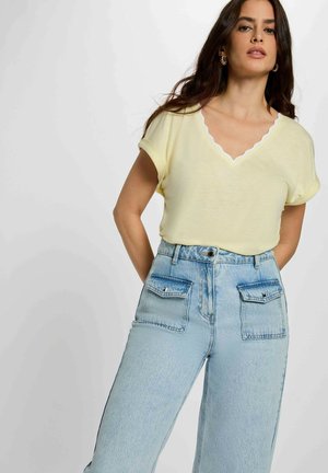 Donna con top giallo chiaro a maniche corte con scollatura a V smerlata e jeans a vita alta azzurri con ampie tasche anteriori.