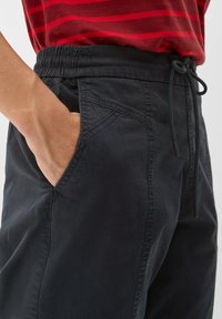 Pantalon à taille élastique noire avec poches latérales, détails à cordon de serrage et texture lisse. Porté avec une chemise rouge à rayures.