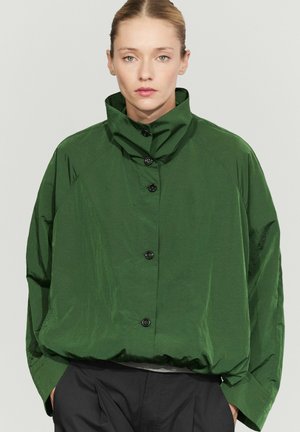 Donna dalla pelle chiara e capelli raccolti indossa una giacca larga verde scuro con collo alto e bottoni e pantaloni neri, in piedi davanti a uno sfondo uniforme.