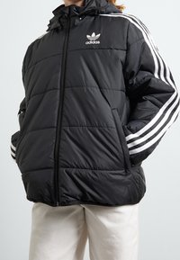 Chaqueta de plumas negra con capucha, con franjas blancas de Adidas en las mangas, cierre de cremallera y bolsillos laterales. Textura suave y acolchada.