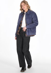 Granatowa kurtka z korduroyu z subtelnym logo kotwicy, nałożona na koszulę w kratkę. Czernie szerokie spodnie i czarne buty dopełniają stylizację.