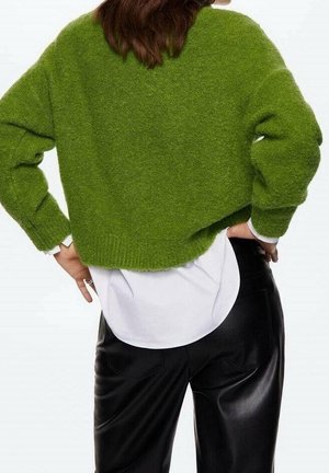 Femme portant un pull court vert duveteux par-dessus une chemise blanche, associé à un pantalon en cuir noir, debout mains sur les hanches, dos face au spectateur.