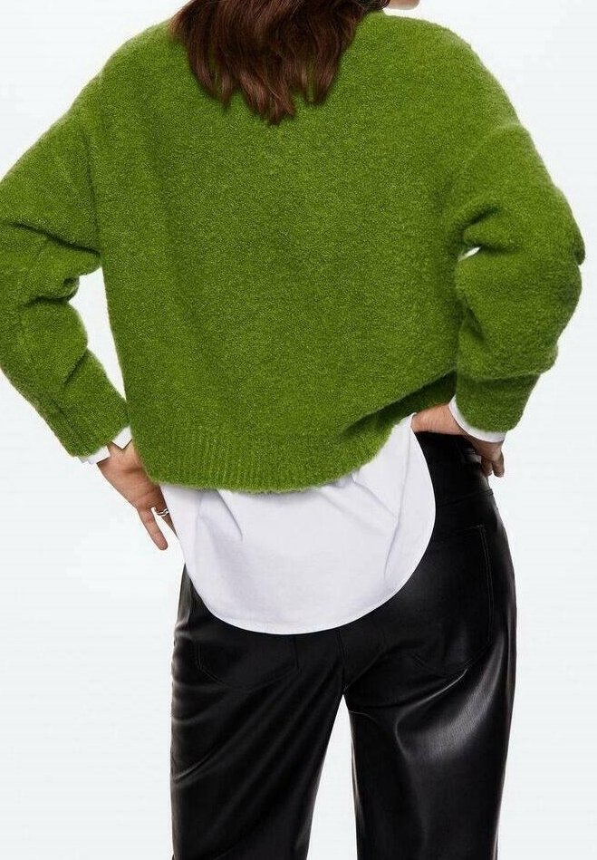 Femme portant un pull court vert duveteux par-dessus une chemise blanche, associé à un pantalon en cuir noir, debout mains sur les hanches, dos face au spectateur.