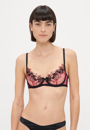 Lacy sheer bra in pink with black floral embroidery, adjustable straps, and a black underband. Bow accent at center.