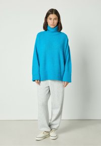 Cianas adīta turtlenecks džemperis ar ribotām aprocēm un apakšmalu, apvienots ar gaiši pelēkiem sporta biksēm un baltām kedām uz vienkrāsaina fona.