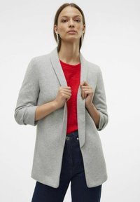 Gilet gris avec un devant ouvert, texture douce, col châle et manches trois-quarts, porté sur un haut rouge et un jean noir.