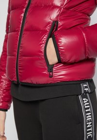 Personne portant une veste rouge matelassée avec la main dans la poche zippée et un pantalon noir avec l'inscription "AUTHENTIC" sur le côté.