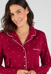 Haut de pyjama rouge avec motif étoilé blanc, passepoil blanc contrastant le long du col et de la poche, et des boutons sur le devant. Tissu doux.