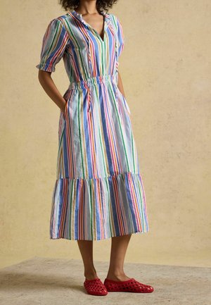 Woman wearing a colorful vertical striped midi dress with puffed sleeves and red woven slip-on shoes standing on a neutral floor.