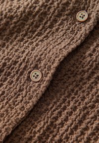 Brauner strukturiierter Stoff mit einem zerknitterten Design und zwei runden Knöpfen. Der Stoff hat eine weiche, leicht raue Textur und ist gefaltet.