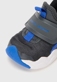 Chaussure de sport noire avec des accents bleus, dotée d'un dessus en tissu texturé, d'une sangle sécurisée et d'une semelle blanche courbée avec une semelle en caoutchouc bleue.