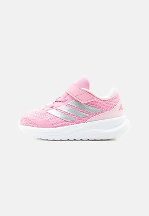Zapatilla deportiva infantil rosa con rayas plateadas, parte superior de malla, cierre de velcro y suela blanca gruesa sobre un fondo claro.