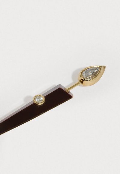 Zlatý sponka do vlasov s dvoma ozdobnými prvkami: hruškovitým falošným drahokamom a okrúhlym čírym kameňom, umiestnenými na tmavo vínovom podklade.
