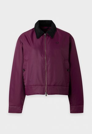Femme debout portant une veste violette, un haut court blanc, une jupe foncée avec des poches, des chaussettes blanches et des mocassins foncés sur un fond uni.
