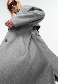 Grijze wollen jas met een relaxte pasvorm, voorzien van een tailleband, knoopsluiting en zijzakken. Model draagt oversized zwarte zonnebril.