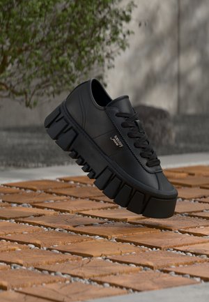 Μαύρο παπούτσι με χοντρή σόλα και λογότυπο "tommy jeans" να αιωρείται πάνω από ξύλινο πλακάκι δαπέδου με πράσινα φύλλα στο φόντο.