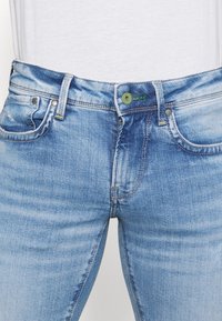 Ljust blå denimjeans med smal passform, med en grön knapp, gula sömmar och två framfickor. Slät textur med blekning.