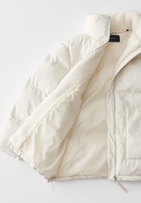 Giacca puffy di colore bianco sporco, con interno foderato in pile e colletto alto. Chiusura con zip e dettagli in tessuto abbinato.